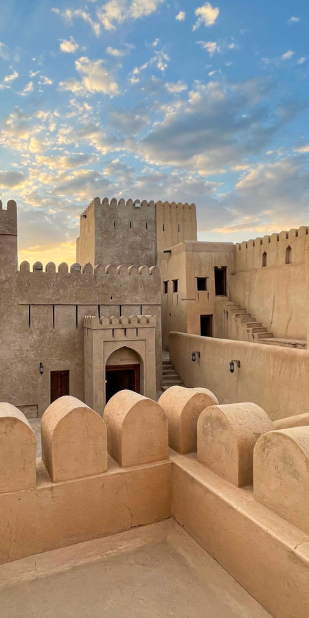 The historic Nizwa Fort in Oman at sunset, its ancient Arabic architecture and defensive walls with towers silhouetted against a dramatic, colorful sky. The fort, built in a traditional Middle Eastern style