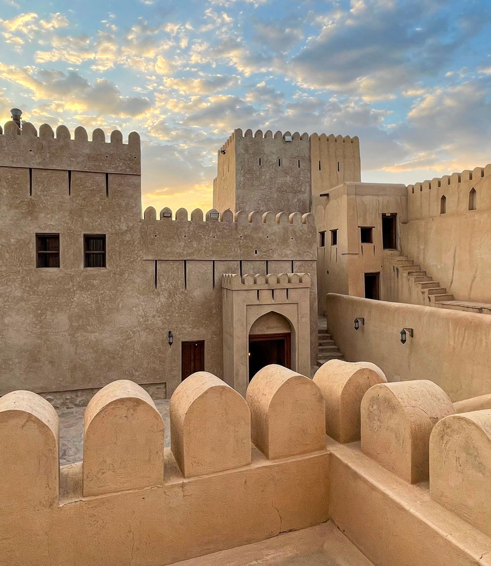 The historic Nizwa Fort in Oman at sunset, its ancient Arabic architecture and defensive walls with towers silhouetted against a dramatic, colorful sky. The fort, built in a traditional Middle Eastern style