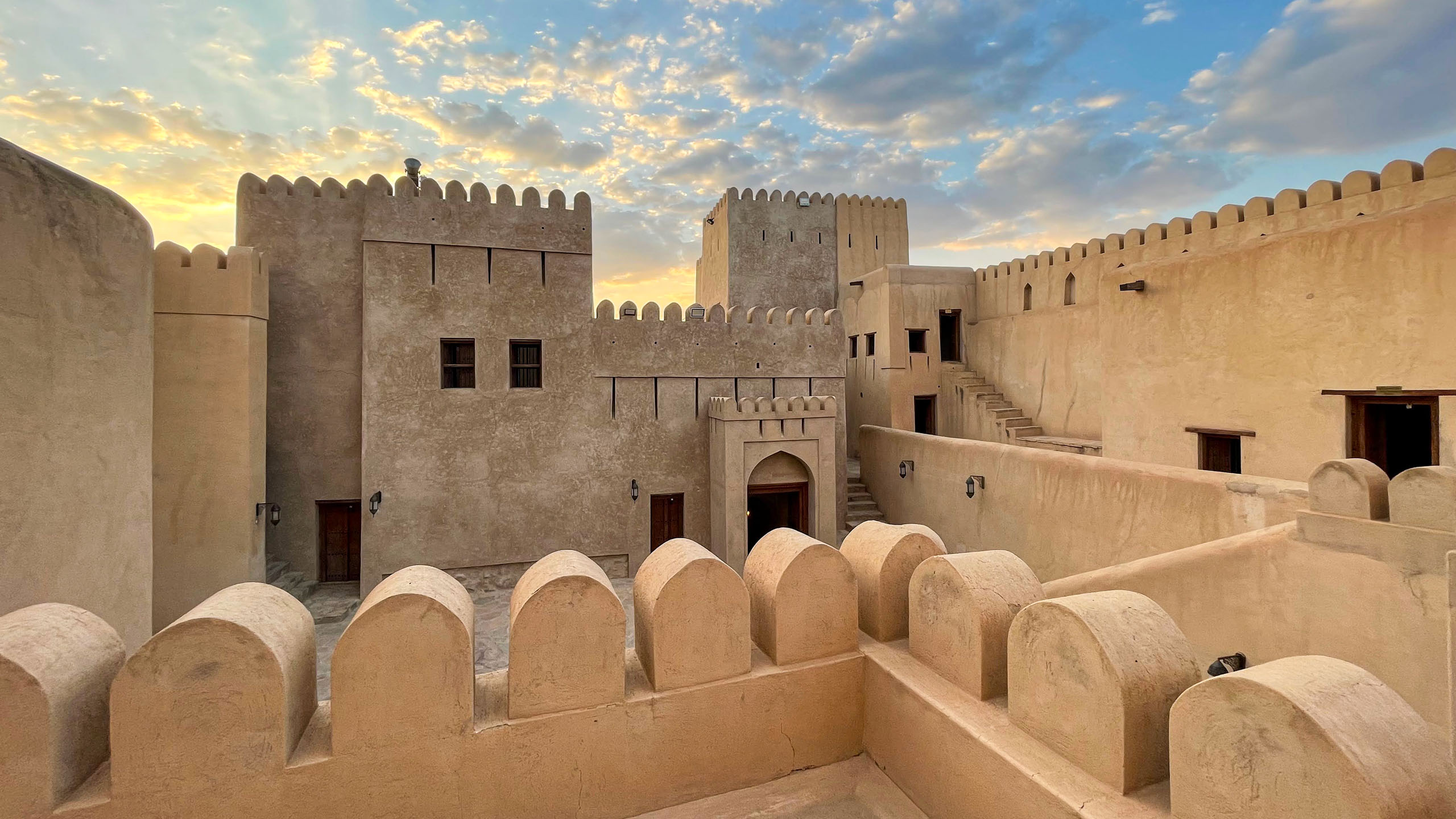 The historic Nizwa Fort in Oman at sunset, its ancient Arabic architecture and defensive walls with towers silhouetted against a dramatic, colorful sky. The fort, built in a traditional Middle Eastern style